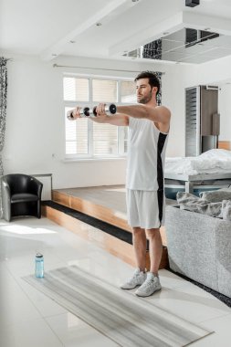 Spor giyimli bir adam spor ayakkabısı giymiş, halterleriyle spor şişesinin yanında egzersiz yapıyor ve evde spor paspası takıyor. 