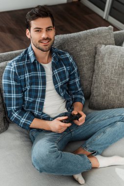 KYIV, UKRAINE - 13 Nisan 2020: Oturma odasındaki kanepede joystick tutarken kameraya gülümseyen yakışıklı adam