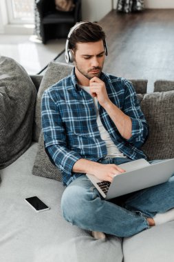 Akıllı telefonun yanında dizüstü bilgisayar kullanan ve kanepesinde boş ekran olan kulaklıklı yakışıklı bir serbest yazar. 