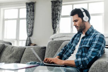 Selective focus of handsome freelancer in headphones working on laptop at home  clipart
