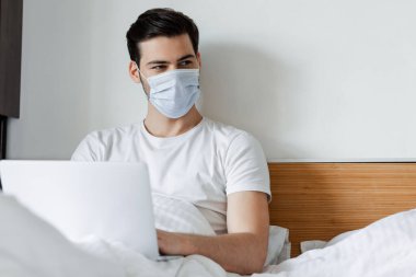 Yatakta dizüstü bilgisayar kullanırken orta maskeli serbest çalışanın seçici odağı. 