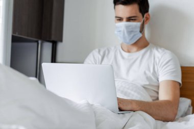 Yatakta dizüstü bilgisayarla çalışan tıp maskeli serbest çalışanın seçici odağı 