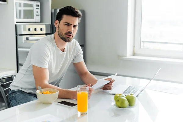 Pensive freelancer looking at camera while holding paper near gadgets and cereals on kitchen table