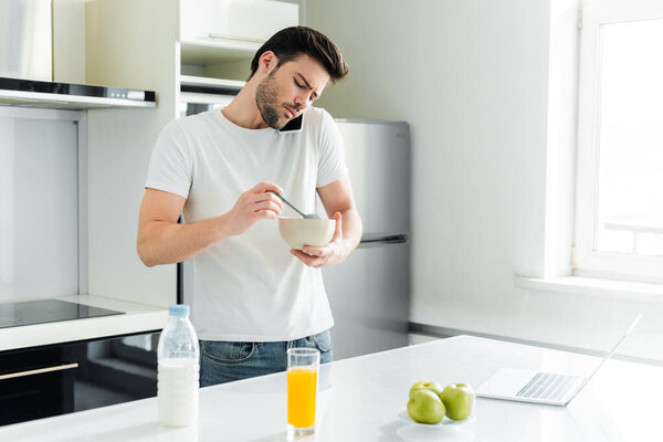 Handsome man talking on smartphone while holding bowl with spoon near laptop and orange juice on kitchen table 
