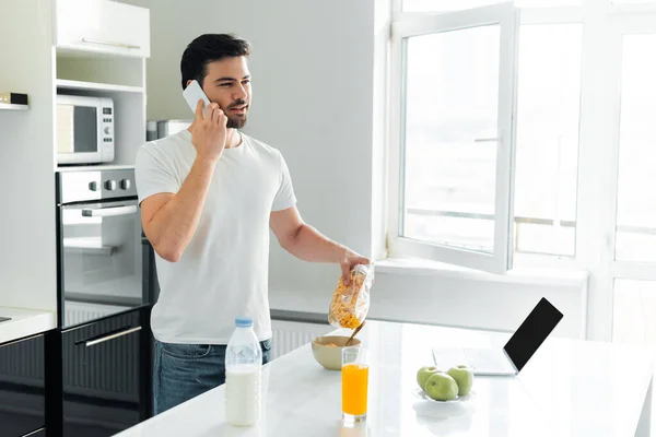 Hombre Hablando Teléfono Inteligente Verter Cereales Tazón Cerca Computadora Portátil —  Fotos de Stock