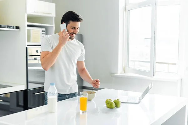 微笑的男人在笔记本电脑旁边用智能手机说话 在厨房桌子上吃早餐 — 图库照片