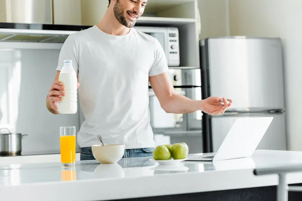 キッチンテーブルのラップトップ上でビデオ通話を持ちながら ミルクのボトルを保持する笑顔の男の選択的な焦点 — ストック写真