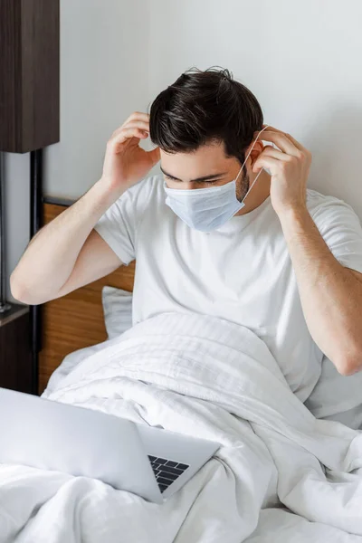 Homem Ajustando Máscara Médica Enquanto Olha Para Laptop Cama — Fotografia de Stock