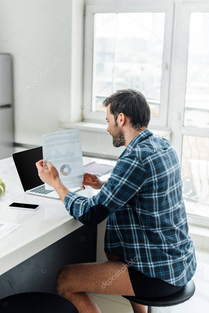 Side view of freelancer in plaited shirt and panties holding paper with charts near laptop and smartphone on table in kitchen 