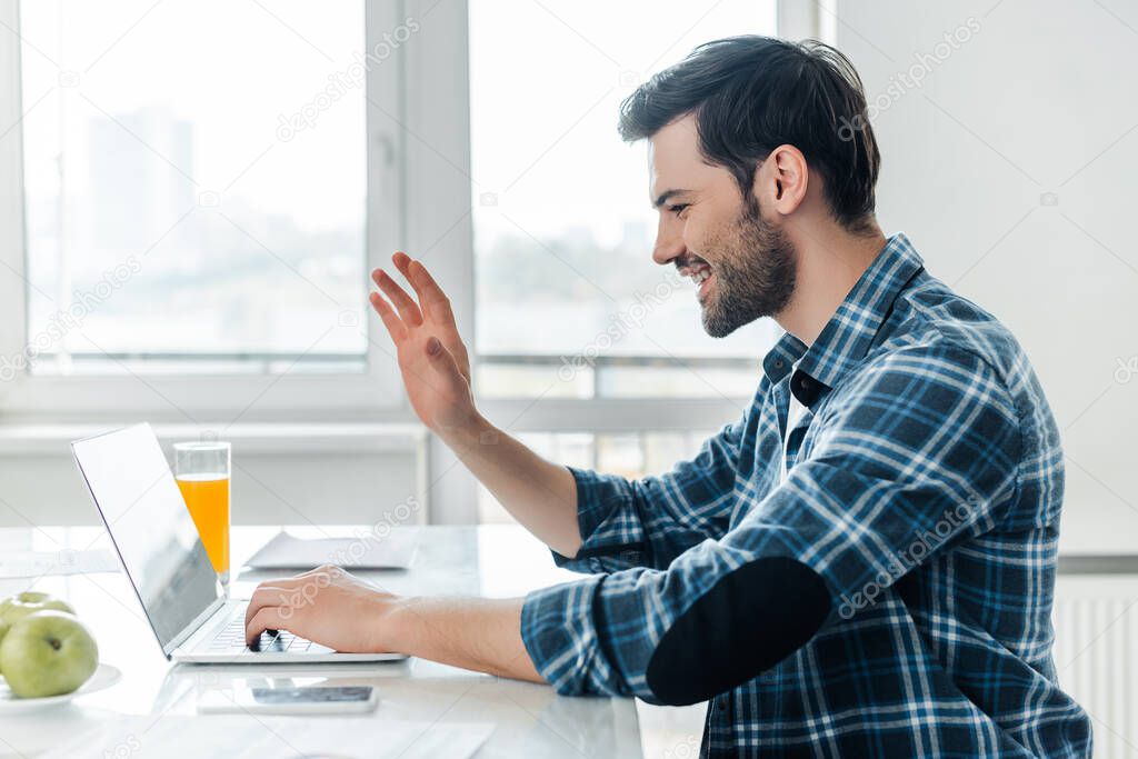 Side view of smiling freelancer having video call on laptop near apples and glass of orange juice on kitchen table 