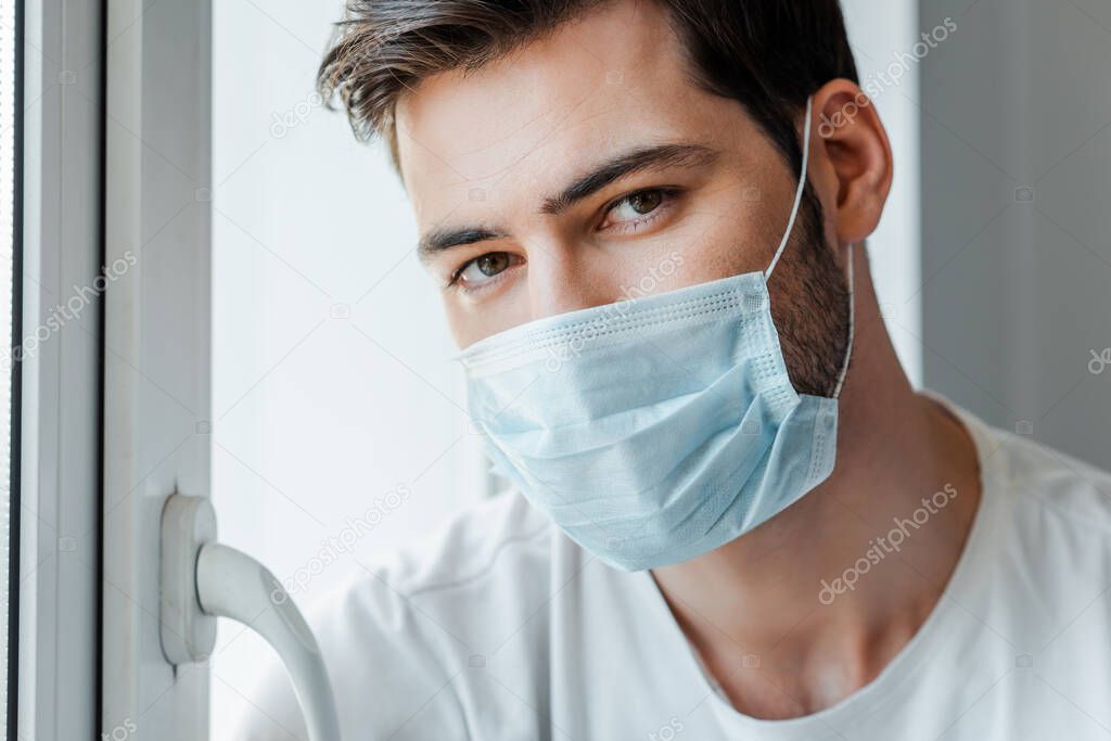 Man in medical mask looking at camera near window at home 