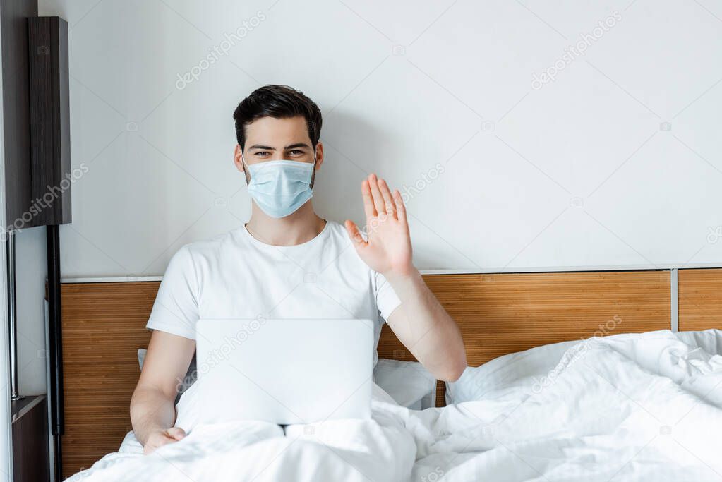 Man in medical mask waving hand at camera while sitting with laptop on bed 