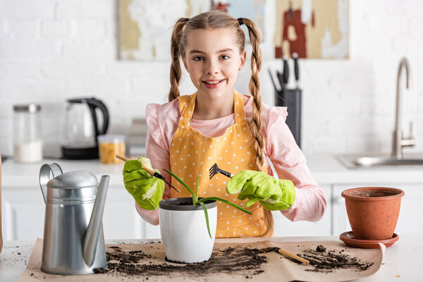Front view of cute child with gardening tools smiling near table with watering pot, ground and flowerpots in kitchen