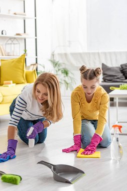 Anne ve kızı ellerinde sprey şişeleri ve paçavralarla oturma odasındaki fırça ve faraş yanında yerleri siliyorlar.