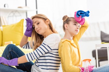 Oturma odasındaki sprey şişesinin yanında sırt sırta oturan bezle yorgun anne ve kız.