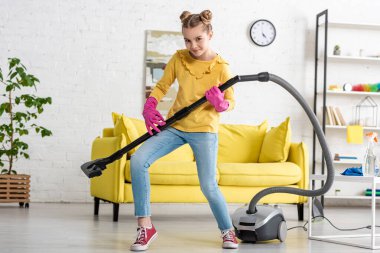 Çocuk elektrikli süpürgeyle gitar çalıyor, gülümsüyor ve oturma odasında kameraya bakıyor.
