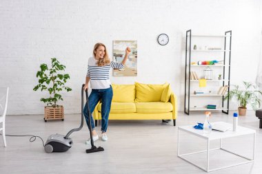 Havalı eli ve elektrikli süpürgesi olan bir kadın oturma odasına bakıyor ve gülümsüyor.