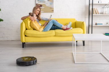 Anne ve kızı dizüstü bilgisayarlarıyla sehpanın yanındaki koltukta dinleniyorlar ve oturma odasının zemininde robotik süpürge var.