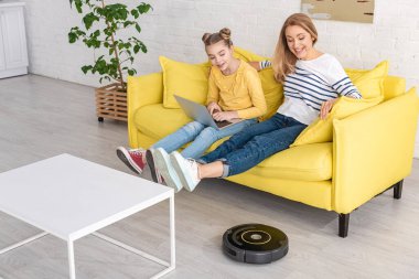Annesinin yanında dizüstü bilgisayarı olan sevimli bir çocuk. Kalkmış bacaklı. Oturma odasındaki kahve masasının yanındaki robot elektrikli süpürgeye bakıyor.