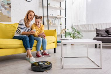 Anne ve kızı, kahve masasının yanındaki kanepede dizüstü bilgisayar ve oturma odasındaki robot süpürgeyle akıllı telefonlara bakıyor.