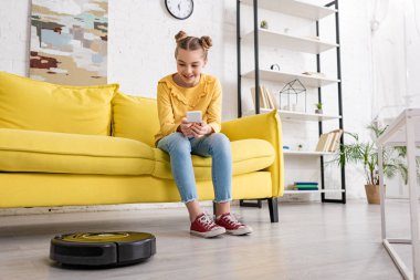 Akıllı telefonlu şirin çocuk kanepede gülümsüyor ve oturma odasında yerde robot süpürgeye bakıyor.