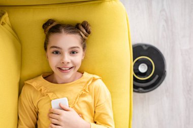 Akıllı telefonla gülümseyen, kameraya bakan ve oturma odasındaki robotik elektrik süpürgesinin yanındaki kanepede uzanan sevimli çocuk manzarası.