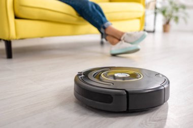 Oturma odasında yerde kadın bacakları ve robotik elektrik süpürgesi var.