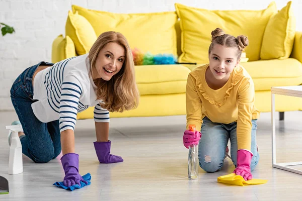 Mother on all fours and daughter with spray bottles and rags wiping floor, smiling and looking at camera in living room