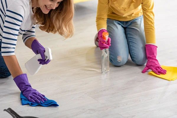 Cropped view of mother and daughter with spray bottles and rags wiping floor in living room