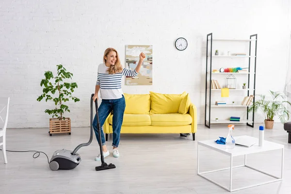 Vrouw Met Opgeheven Hand Stofzuiger Die Wegkijkt Glimlacht Woonkamer — Stockfoto