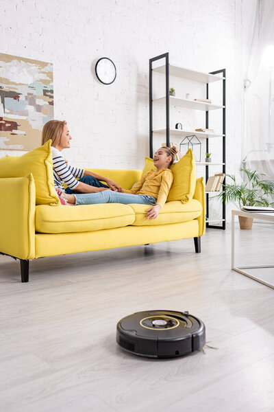 Mother and daughter looking at each other and lying on sofa near coffee table and robotic vacuum cleaner on floor in living room