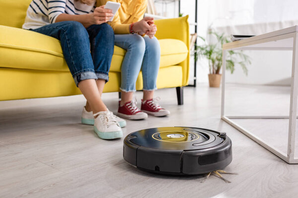 Cropped view of woman with smartphone and daughter on sofa near coffee table and robotic vacuum cleaner on floor in living room