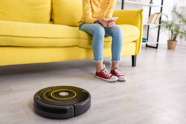 Partial view of child with smartphone on sofa near robotic vacuum cleaner on floor in living room