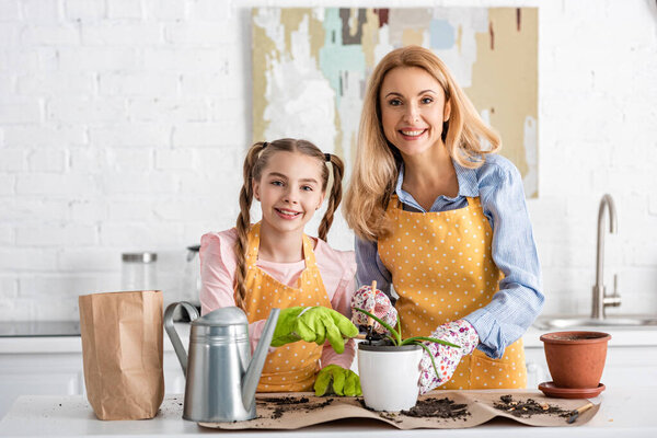 Mother and cute daughter smiling with gardening tools and flowerpot with aloe near watering pot and paper bag with ground on table 