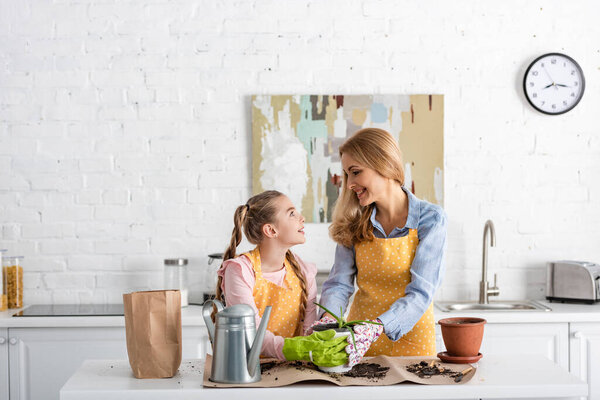 Mother and cute daughter looking at each other and touching flowerpot with aloe near watering pot and paper bag on table with ground