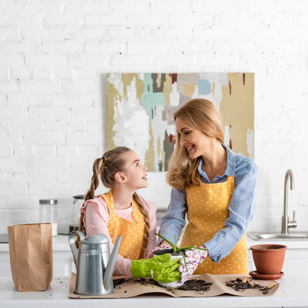 Mother and daughter looking at each other and touching flowerpot with aloe near watering pot and paper bag on table with ground