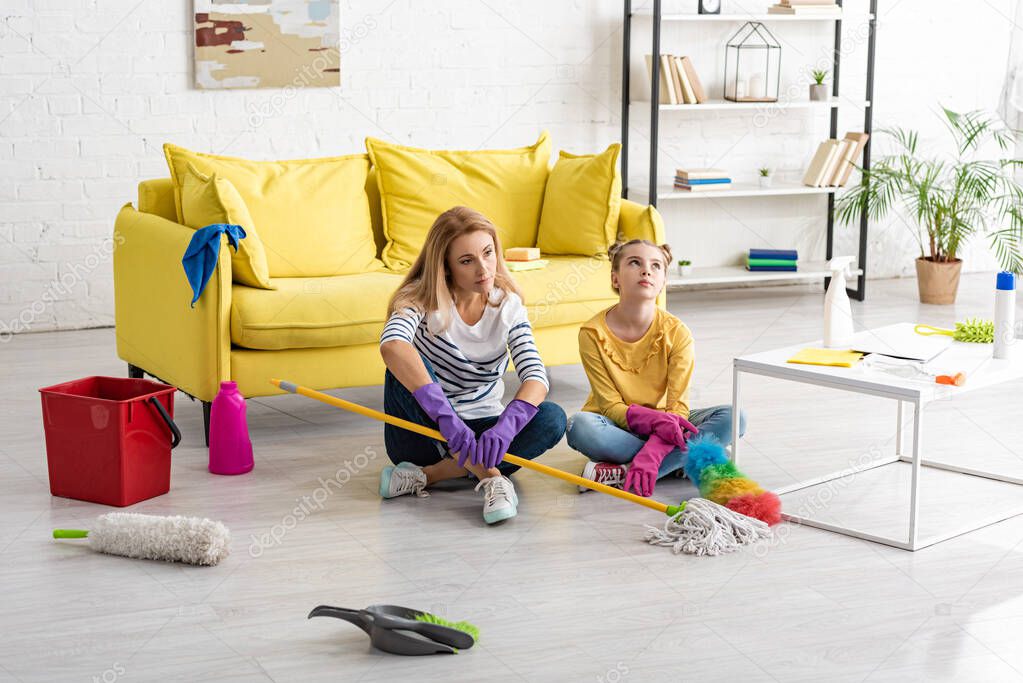 Tired mother and daughter with crossed legs and cleaning supplies on floor near coffee table in living room