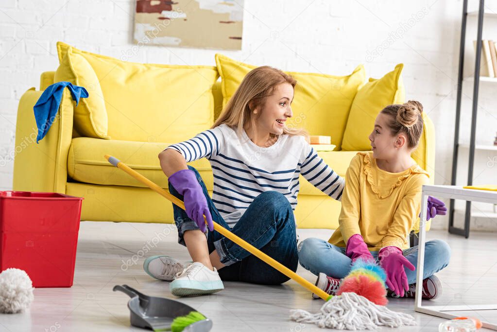 Surprised mother and daughter with crossed legs and cleaning supplies looking at each other on floor in living room