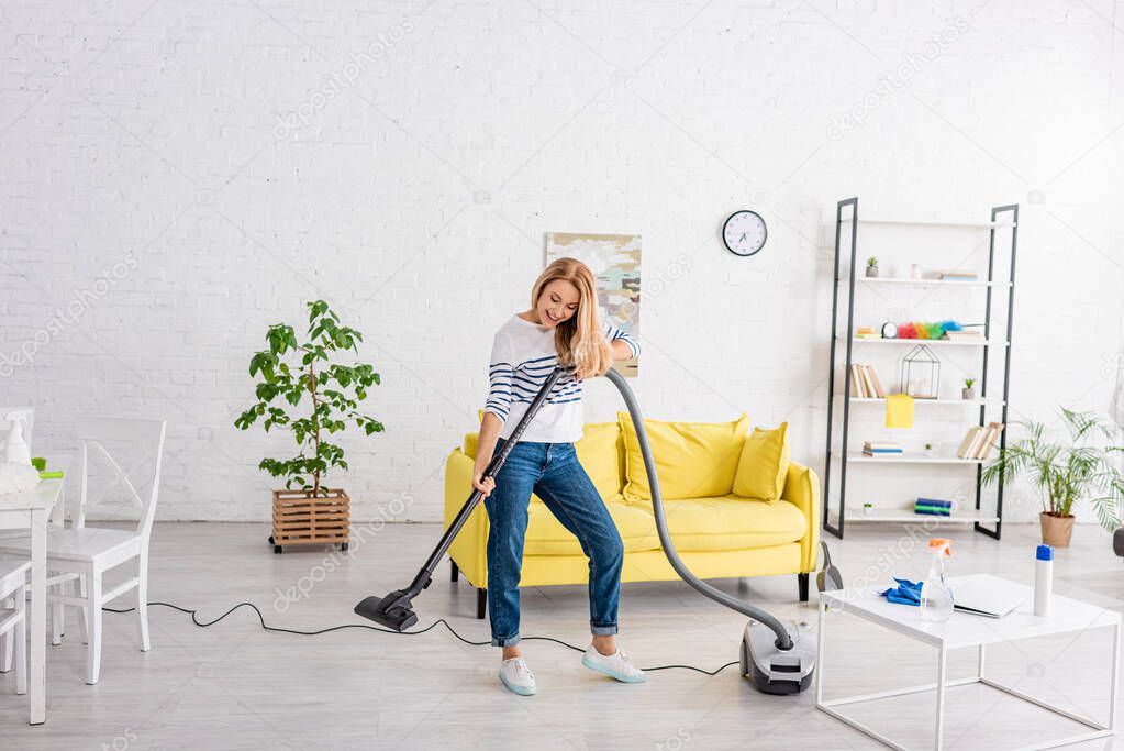 Blonde woman smiling and cleaning up with vacuum cleaner in living room