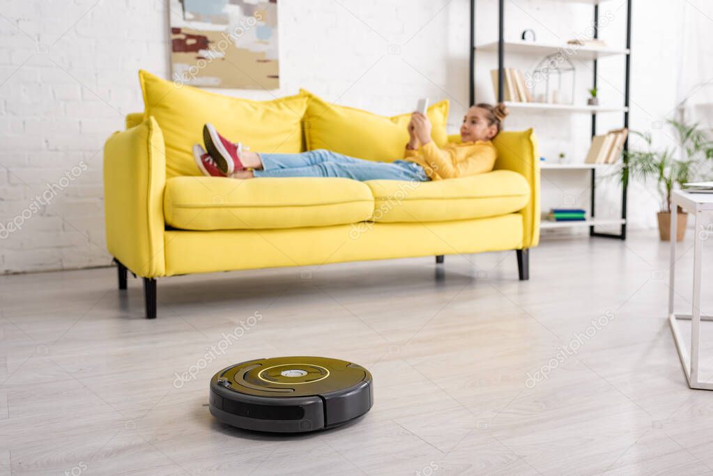 Selective focus of kid with smartphone lying on sofa near robotic vacuum cleaner on floor in living room