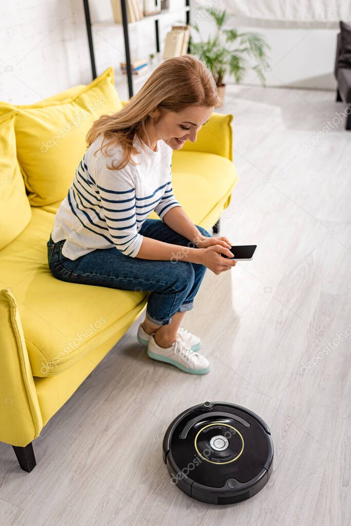 High angle view of woman smiling with smartphone on sofa near robotic vacuum cleaner on floor in living room