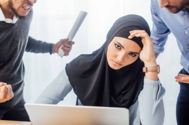 Kızgın Müslüman iş kadınının odak noktası tartışan meslektaşlarının yanındaki kameraya bakıyor. 