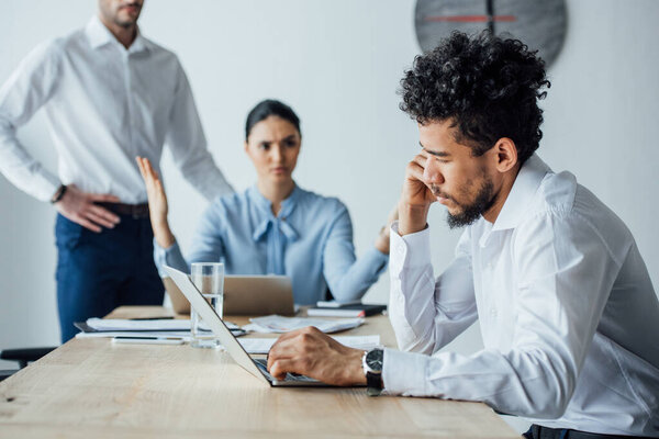 Селективный фокус африканского американца, использующего ноутбук рядом с сердитым мексиканским коллегой в офисе
 