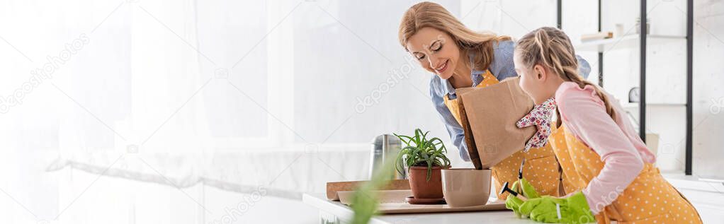 Horizontal image of happy mother with paper bag putting ground to flowerpot and cute daughter with rake near table in kitchen