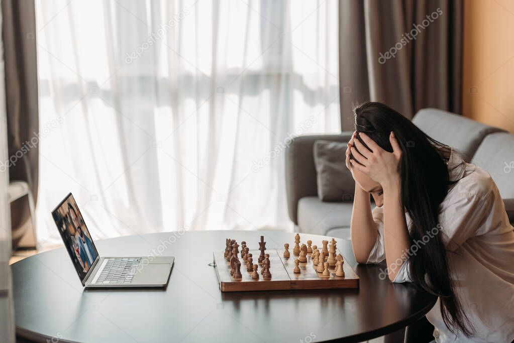 depressed woman touching bowed head while having video chat with boyfriend near chessboard