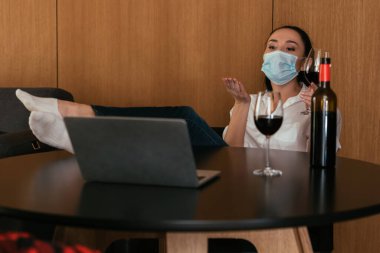 Tıbbi maskeli genç bir kadın görüntülü konuşma sırasında şişe ve kırmızı şarap kadehlerinin yanında hava öpücüğü gönderiyor.