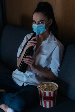 Sağlık maskeli depresif kadın elinde şarap şişesiyle patlamış mısır kovasının yanındaki kameraya bakıyor.