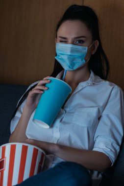 Elinde soda ve patlamış mısır tutarken kameraya göz kırpan tıbbi maskeli genç bir kadın. 