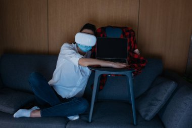 VR kulaklıklı genç bir kadın koltukta oturuyor ve dizüstü bilgisayarına sarılıyor.