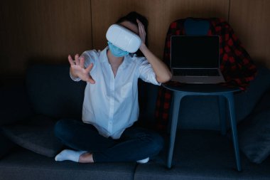 Bilgisayarın yanındaki koltukta boş bir ekranla otururken, uzanmış eli kulaklık kullanan genç bir kadın.